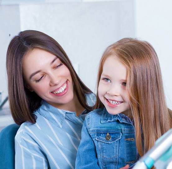 Girl in a jean jacket smiling up at her orthodontist from her mother’s lap